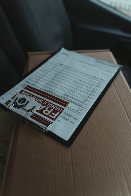 A close-up of a packing list document placed on top of a cardboard box inside a moving van, illustrating the home relocation process. The document, secured with a clear plastic cover displaying a prom